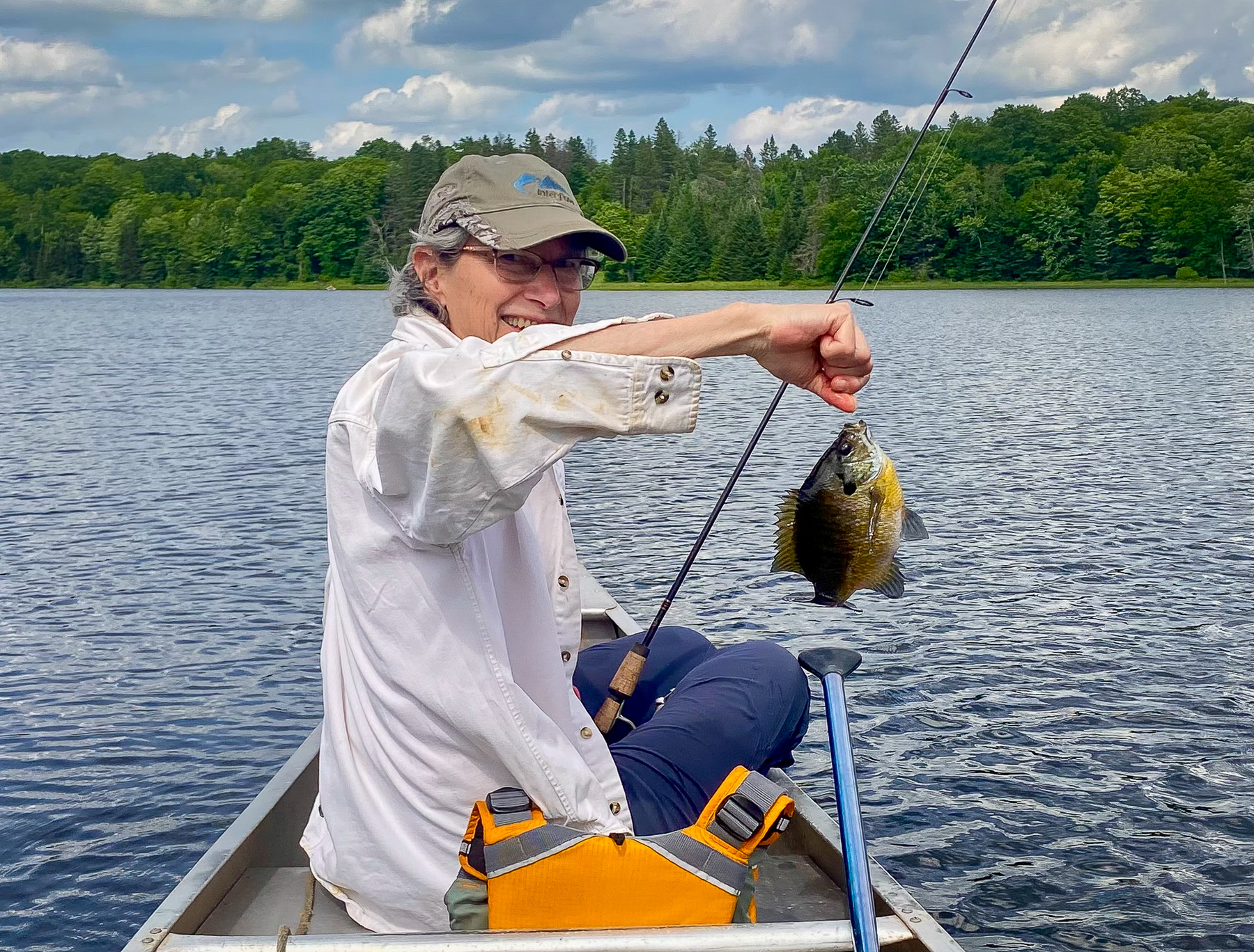 Person in a white shirt and cap on a small boat holds a caught bass on a fishing line over a lake; trees and clouds behind.